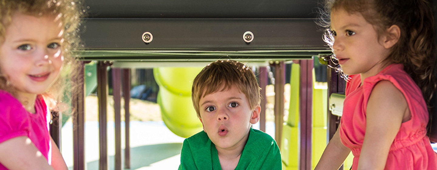 Three kids gather in the shade underneath a playground at a Table Panel.