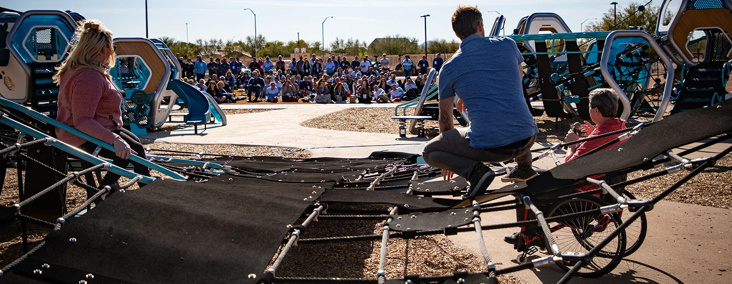 Image taken from inside of a net structure. There are three speakers that are speaking to a crowd of people. Image is taken from behind speakers. There are two Hedra play structures on either side of the speakers.