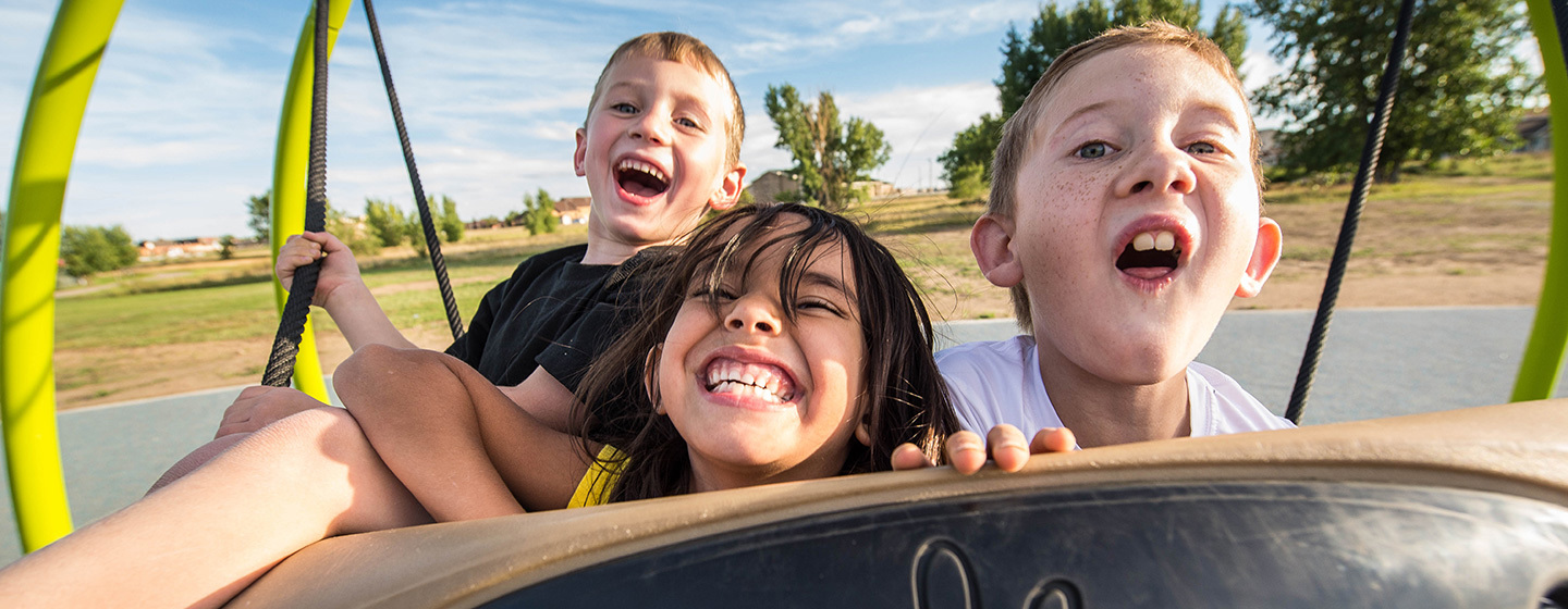 Three kids smiling while they swing together on an inclusive playground Oodle Swing.