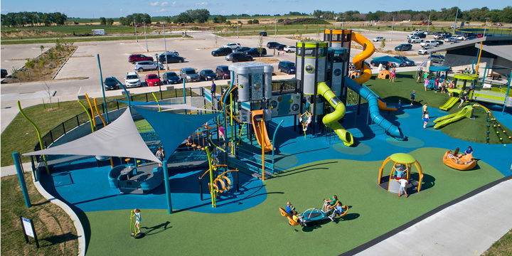 Aerial view of a playground with three large silver towers resembling spaceships with multiple freestanding play elements around and triangular shades to the left. 