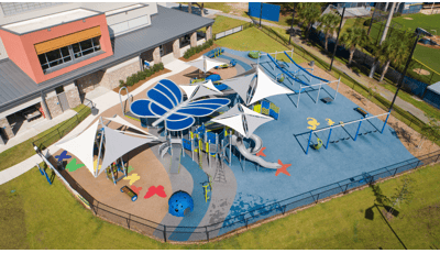 Aerial view of playground with a blue butterfly shaped shade structure. Playground surfacing includes colorful butterflies and starfish all next to school building. 